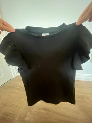 Svart topp med volangärm - Snygg svart topp med volang på ärmarna. Toppen har en klassisk rund halsringning och är tillverkad i ett mjukt material som sitter bekvämt. Perfekt för dig som gillar en enkel men trendig stil.