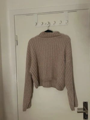 Beige ribbstickad tröja från Gina Tricot - Mysig beige ribbstickad tröja från Gina Tricot med bred krage och långa ärmar. Tröjan har en avslappnad passform och är tillverkad i ett mjukt, chenille-liknande material som känns skönt mot huden. Perfekt för lager-på-lager-stil.