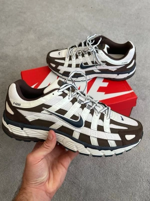 Nike P-6000 bruna och vita sneakers - Nike P-6000 sneakers i brunt och vitt med mesh och syntetiska överdrag. Skorna har en sportig retrodesign, tydlig Swoosh-logga på sidan och tjock sula för extra komfort. Perfekta för dig som gillar streetwear och vill sticka ut med en unik look.