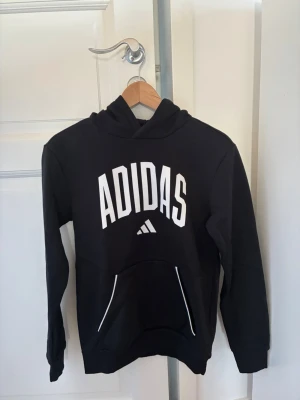 Svart hoodie från Adidas med tryck - Snygg svart hoodie från Adidas med stor vit logga. Klassisk känguruficka framtill och huva. Prislappen är borttagen men tröjan är aldrig använd. 