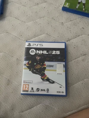 NHL 25 till PS5 - NHL 25 till PlayStation 5, ett ishockeyspel från EA Sports. Omslaget är blått och vitt med en hockeyspelare i svart och gult på framsidan. Spelet levereras i ett klassiskt PS5-fodral av plast och inkluderar skiva samt bonuskod.
