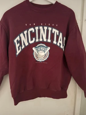Vinröd sweatshirt Pull&Bear XS - Cool vinröd sweatshirt från Pull&Bear med tryck 'ENCINITAS San Diego' och basebollmotiv på bröstet. Oversized passform, ribbade muddar vid hals, ärmslut och nederkant. Perfekt för en avslappnad streetstyle-look.