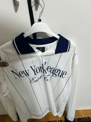 Vit randig långärmad skjorta H&M storlek S - Vit långärmad skjorta från H&M med blå och gröna ränder samt mörkblå krage. Tryck med texten 'New York League East Est.' framtill. Skjortan har knappar vid halsen och sportig känsla.