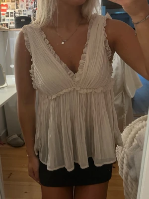 Ljusvit/biehe volangblus med v-ringning - Supersöt blus i ljus vit/biege färg med djup v-ringning och volangdetaljer längs kanterna. Plisserad och luftig modell som ger en romantisk vibe. Perfekt till sommarens outfits och lätt att matcha med både kjol och jeans.