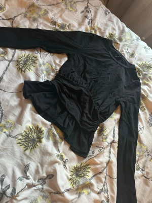 Svart top från Gina Tricot - Säljer en svart omlottopp från Gina Tricot i mjukt bomullsmaterial. Toppen har långa ärmar och en snygg volangdetalj framtill som ger en extra touch till outfiten. Perfekt att styla med jeans eller kjol.