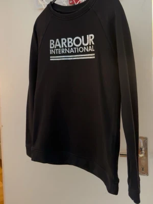 Svart sweatshirt från Barbour - Svart sweatshirt från Barbour International med vit logga tryckt på bröstet. Tröjan har rund halsringning och långa ärmar. Tillverkad i mjukt material som passar perfekt till en avslappnad stil.