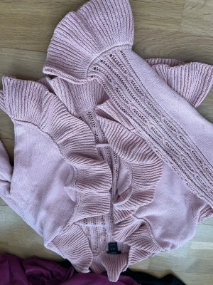 Rosa volangstickad kofta från Lindex - Supersöt ljusrosa stickad kofta från Lindex med volangdetaljer längs kanterna och ribbstickade muddar. Koftan har ett öppet hålmönster framtill och är perfekt för dig som gillar romantisk och trendig stil.