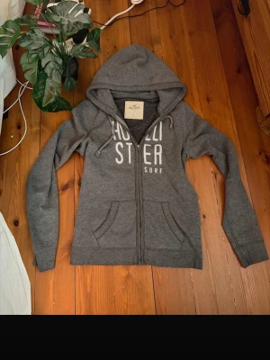 Grå Hollister hoodie med dragkedja - Galet snygg hoodie från Hollister med vit text på bröstet och dragkedja framtill. Tröjan har huva med dragsko och två fickor fram. Perfekt för chill dagar och helt perfekt nu för hösten, tröjan är i storlek L men sitter som m/s 