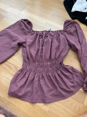 Lila offshoulder topp med puffärm - Trendig lila topp med offshoulder-design och puffiga långärmar. Smockad midja ger en snygg siluett och toppen har en lätt rynkad struktur. Perfekt för dig som vill ha en feminin och modern look.