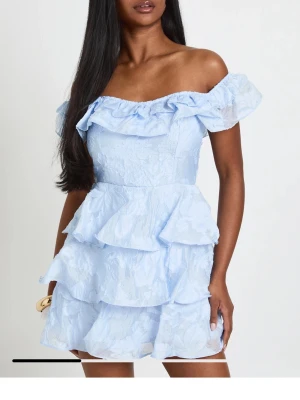 Volangklänning - Supersöt ljusblå kortklänning med offshoulder och flera lager volanger. Klänningen har en romantisk look med rynkade detaljer och axelbandslös design. Perfekt för sommarens alla tillfällen. 