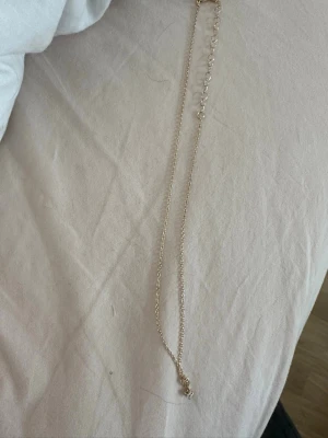 Guldigt halsband med klar sten - Snyggt halsband i guldfärgad metall med en tunn kedja och ett hängsmycke med en klar sten. Perfekt för att ge din outfit lite extra bling. Låset är justerbart så du kan anpassa längden efter din stil.