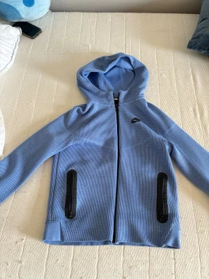 Nike Thec  - Säljer en ljusblå Nike hoodie för barn  med dragkedja framtill och två svarta dragkedjeförsedda fickor. Hoodien har en sportig look med ett diskret rutmönster i tyget och huva. Perfekt för chill dagar eller när du vill ha en avslappnad stil.