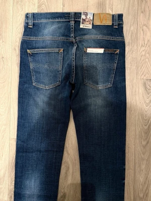 Helt Nya Nudie Jeans Thin Finn Blå W30/L34 - Slim Fit - Nudie Jeans Thin Finn ”Indigo Dreams” blå jeans med slim fit-passform, låg midja och avsmalnande ben, storlek W30/L34, midja 38,5 cm, ytterbenslängd 111 cm, benöppning 16 cm, modellen är 185cm & väger 65kg, Helt nya med tags.
