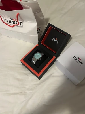 Tissot silverfärgad klocka - Snygg klocka från Tissot med silverfärgat armband i metall och ljus urtavla. Klockan har en klassisk rund form och levereras i originalbox med garantikort och manual. Perfekt accessoar för dig som gillar stilrena detaljer.