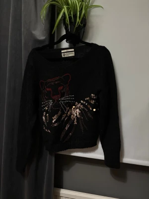 Svart stickad tröja med paljetter - Cool svart stickad tröja med ett broderat pantermotiv framtill och detaljer i guldiga paljetter. Tröjan har rund halsringning och långa ärmar. Perfekt för dig som vill sticka ut lite extra med ett djurmotiv och glittriga detaljer.