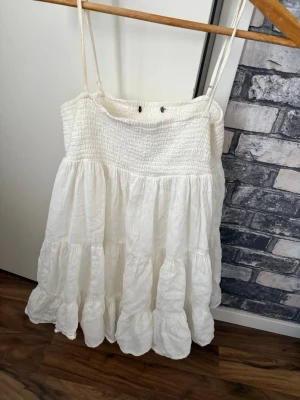 Vit sommarklänning med volanger - Luftig vit klänning med smala axelband och volangdetaljer. Klänningen har en smockad överdel och är tillverkad i ett lätt material, perfekt för varma dagar. Passformen är loose och modellen är kortklänning.
