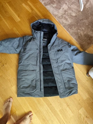 Grå dunjacka med huva från Helly Hansen - Snygg grå dunjacka från Helly Hansen med svart HH-logga på bröstet och ryggen. Jackan har huva, dragkedja och stora fickor framtill. Insidan är svart och quiltad för extra värme. Perfekt för kalla vinterdagar och riktigt skön passform.