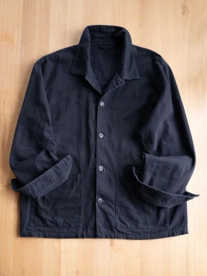 Black Chore Jacket, size XL - Säljer en svart overshirt i kraftigt bomullstyg med klassisk krage och fyra knappar framtill. Två stora fickor framtill och uppvikta ärmar ger en avslappnad look. Perfekt lager på lager-plagg för dig som gillar minimalistisk stil.