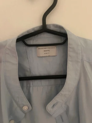 Ljusblå skjorta från Selected Homme - Säljer en ljusblå skjorta från Selected Homme med ståkrage och knappar framtill. Skjortan har lång ärm och är i slim fit-modell, perfekt för en clean och stilren look. Tillverkad i mjuk bomull för skön känsla.