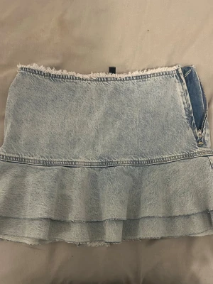 Ljusblå jeanskjol med volang - Trendig ljusblå jeanskjol med råa kanter och volang nertill. Kjolen har en dragkedja i sidan och är tillverkad i denim med en kort passform. Perfekt för dig som vill ha en cool och avslappnad stil.