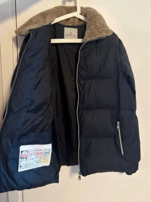Svart dunjacka med teddykrage Moncler - Snygg blå dunjacka från Moncler med dragkedja och en mysig beige teddykrage. Jackan har en innerficka med serietryck, quiltad design och en ficka med dragkedja på sidan. Perfekt för kalla vinterdagar.