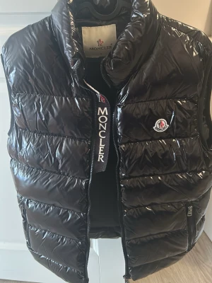 Svart dunväst från Moncler - Snygg svart dunväst från Moncler med glansig finish och hög krage. Västen har dragkedja framtill, två sidofickor med dragkedja och Moncler-logga på bröstet. Perfekt för lager på lager och streetstyle. VÄSTEN ÄR TROLIGTVIS INTE ÄKTA!!!!! 