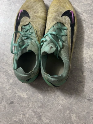Nike Phantom GT fotbollsskor mint/gul - Säljer ett par Nike Phantom GT fotbollsskor i mintgrönt och gult med svarta och lila detaljer. Skorna har snörning, låg profil och är tillverkade i syntetmaterial för bra bollkontroll. Perfekta för dig som vill sticka ut på planen.