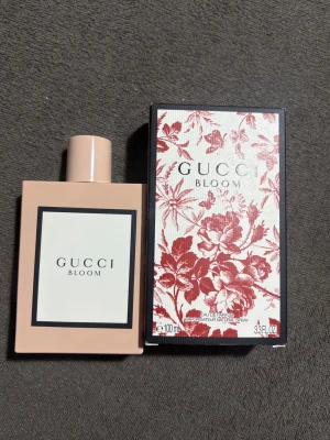 Gucci Bloom Eau de Parfum 100 ml - Gucci Bloom Eau de Parfum i en stilren, rektangulär ljusrosa flaska med matchande lock. Förpackningen har ett elegant blommönster i rött och vitt. Doften är känd för sina blommiga och fräscha noter, perfekt för dig som gillar exklusiva parfymer.