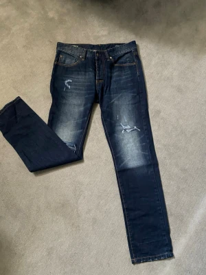 Mörkblå straight fit jeans CKH Denimite - Snygga mörkblå jeans från Clockhouse, modell CKH Denimite. Straight fit med slitningar och ljusare partier på låren och knäna för en trendig look. Klassisk femficksmodell med orange sömmar. Perfekta för dig som gillar en avslappnad stil.