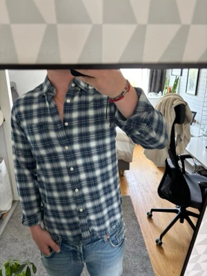 Flanell - Säljer en blå och vit rutig skjorta med långa ärmar och klassisk krage. Skjortan har knäppning framtill och en bröstficka. Perfekt att bära öppen över en t-shirt för en avslappnad stil.
