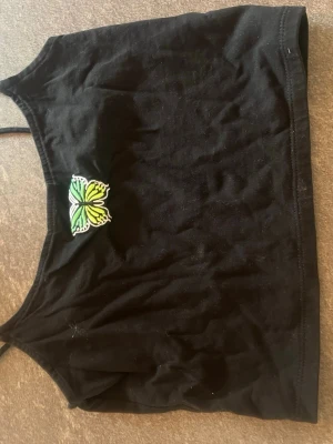 Svart croppad linnetopp med fjäril - Svart croppad linnetopp från FB Sister med smala axelband och en broderad grön och gul fjäril framtill. Perfekt för varma dagar och enkel att matcha med jeans eller kjol.