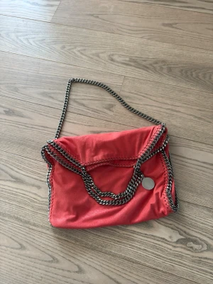 Röd Stella McCartney axelväska - Cool röd/korall axelväska från Stella McCartney med kedjedetaljer i silver runt kanterna och som axelrem. Väskan har en mjuk, slouchy form och är tillverkad i veganskt läder. Rund metallbricka med logga hänger på sidan för extra detalj. Väskan är köpt på Vestiaire för ca 4500 och är har knappt använts sedan inköp.