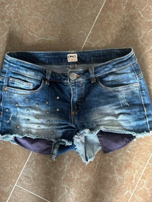 Blå jeansshorts med stjärndetaljer ONLY - Säljer ett par blå jeansshorts från ONLY med slitna kanter och stjärnformade nitar på ena sidan. Shortsen har fem fickor, knappgylf och klassisk denim-look med ljusa slitningar. Perfekta för sommaren!