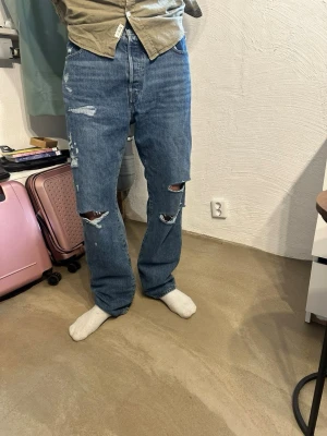 Levis 501 jeans - Säljer ett par blå raka jeans med stora slitningar och hål på båda benen. Jeansen har klassisk femficksdesign och hög midja. Materialet är kraftig denim i bomull och passformen är avslappnad och loose fit.