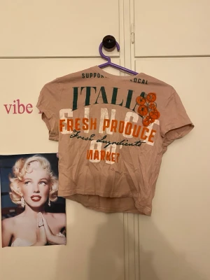 Beige croppad t-shirt med tryck - Säljer en beige croppad t-shirt med färgglatt tryck på framsidan där det står 'ITALIAN FRESH PRODUCE MARKET' och illustration av apelsiner. T-shirten har korta ärmar och en avslappnad passform. Perfekt för dig som gillar unika prints och streetstyle.