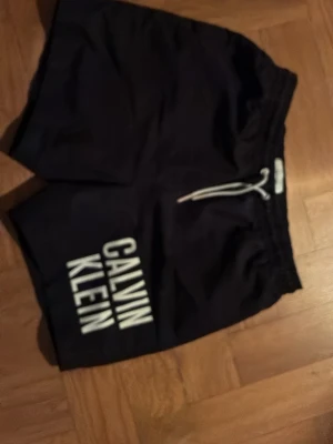 Svarta badbyxor från Calvin Klein - Säljer ett par svarta badbyxor från Calvin Klein med vit logga tryckt längs ena benet. De har resår i midjan och vit dragsko för justerbar passform. Perfekta för stranden eller poolen.