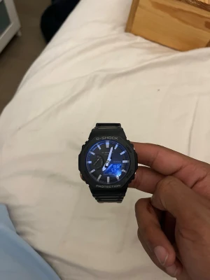 Svart G-Shock digital klocka - Säljer en svart G-Shock klocka med digital och analog display. Klockan har en robust design med tydliga vita markeringar och blå bakgrundsbelysning. Armbandet är i slitstark syntet och boetten är rund med skyddande kanter. Perfekt för dig som gillar sportig och tålig stil.