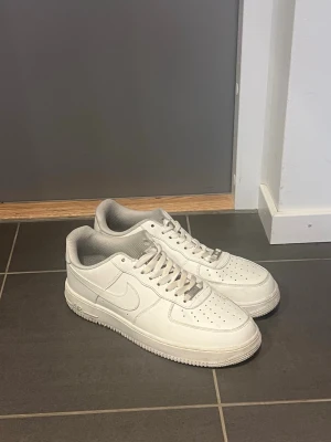 Nike Air Force 1 sneakers - Klassiska vita Nike Air Force 1 sneakers i lågt utförande. Skorna har en stilren design med perforerad tå, vita skosnören och diskret Nike-logga på sidan. Tillverkade i läder med en bekväm och vadderad insida. Perfekta för en clean och tidlös look.
