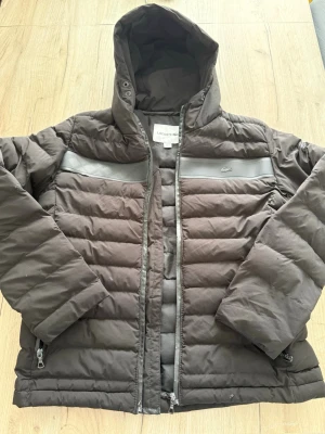 Svart pufferjacka från Lacoste - Säljer en svart pufferjacka från Lacoste med huva och dragkedja. Jackan har diskreta grå detaljer över bröstet och två fickor med dragkedja. Klassisk Lacoste-logga på bröstet. Perfekt för kalla vinterdagar.