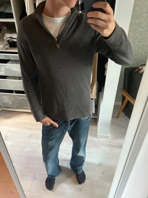 Mörkgrå halvzip tröja Polo Ralph Lauren - Mörkgrå långärmad tröja från Polo Ralph Lauren med halvzip och liten broderad logga på bröstet. Tröjan har en hög krage och är tillverkad i mjukt material som känns skönt mot huden. Perfekt för en avslappnad och stilren look.