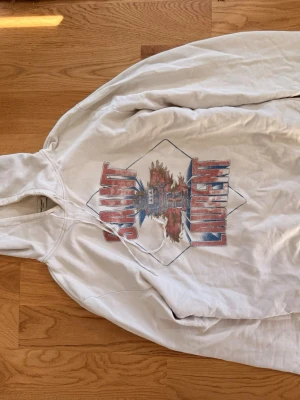 Vit hoodie från Saint Laurent - Säljer en vit hoodie från Saint Laurent med tryck i rött och blått framtill där det står 'Saint Laurent' och ett grafiskt motiv. Hoodien har huva och är tillverkad i mjukt bomullsmaterial. Perfekt för en avslappnad och trendig look.