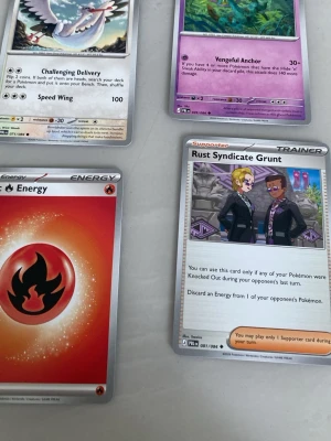 Pokémonkort blandade typer - Fyra Pokémonkort: ett med eldenergi, ett tränarkort 'Rust Syndicate Grunt', samt två Pokémonkort med olika motiv och färger. Korten har klassisk rektangulär form och är tryckta på glansigt papper. Perfekt för samlare eller Pokémon TCG-spelare.