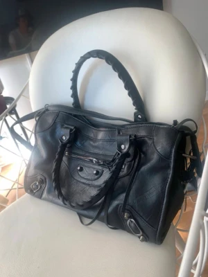 Svart väska💓 - Snygg svart handväska i mjukt läder med flätade handtag och silverfärgade detaljer. Väskan har en framficka med dragkedja och dekorativa nitar. Perfekt storlek för att få plats med allt du behöver och ger en cool vibe till din outfit.💓