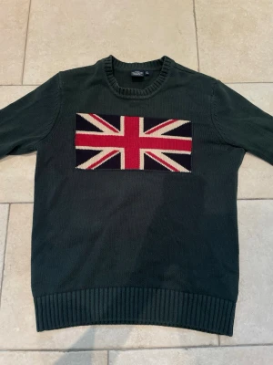Mörkgrön stickad tröja Crocker - Säljer en mörkgrön stickad tröja från Crocker med Union Jack-flagga i rött, vitt och blått på bröstet. Tröjan har rund halsringning, ribbade muddar och nederkant. Perfekt för dig som gillar klassisk brittisk stil och vill sticka ut.