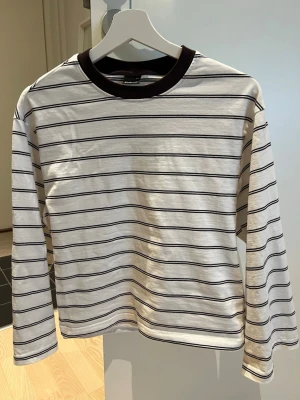 Randig långärmad topp Gina Tricot - Säljer en vit långärmad topp från Gina Tricot med tunna bruna ränder och kontrasterande brun krage. Perfekt till jeans eller kjol för en enkel och stilren look. Tillverkad i mjuk bomull som känns skön mot huden.