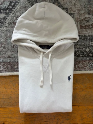 Vit hoodie från Polo Ralph Lauren - Snygg vit hoodie från Polo Ralph Lauren med klassisk broderad logga i marinblått på bröstet. Hoodien har dragsko i huvan, känguruficka och ribbade muddar vid ärmar och nederkant. Tillverkad i mjuk bomull för maximal komfort. OBSERVERA❗️Tröjan har tyvärr ett par mindre fläckar som syns på bilderna. Det kan förekomma någon mer prick/fläck som jag missat. Med hänsyn till detta är priset nedsatt med ca 200kr.