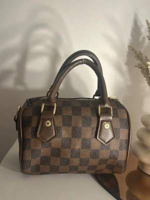 Brun rutig handväska i läderlook - Snygg handväska i brun och mörkbrun rutig design med guldfärgade detaljer. Väskan har två stadiga handtag och dragkedja upptill. Klassisk boxig form och stilren look, perfekt för att lyfta vilken outfit som helst.