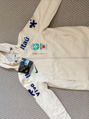  Brasilien Nike windbreaker  - Snygg vit vindjacka från Nike med Brasilien-logga och sponsormärken som Itaú och Vivo. Jackan har huva, dragkedja framtill, två sidofickor och är tillverkad i GORE-TEX-material. Perfekt för dig som gillar sportig stil och vill sticka ut.