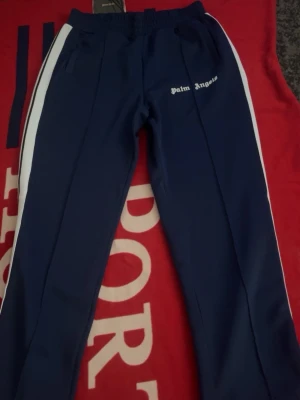 Blå  Palm Angels mjukisbyxor - Snygga blåa mjukisbyxor från Palm Angels med vita sidoränder och logga på vänster lår. Byxorna har resår i midjan och raka ben. Perfekta för en chill och trendig look. Pris kan diskuteras och mer bilder kan skickas 