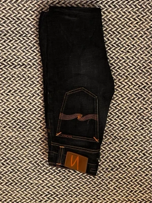 Mörkblå jeans från Nudie Jeans - Säljer ett par mörkblå jeans från Nudie Jeans med klassisk orange kontrastsöm och vågig detalj på bakfickan. Jeansen har fem fickor, raka ben och är tillverkade i kraftig denim. Perfekta för dig som gillar stilrena och tidlösa jeans.